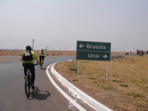 Imagem mostra ciclistas fazendo o contorno do balão, seguindo sentido Unaí, com a placa da divisa, Brasília / Unaí à sua direita