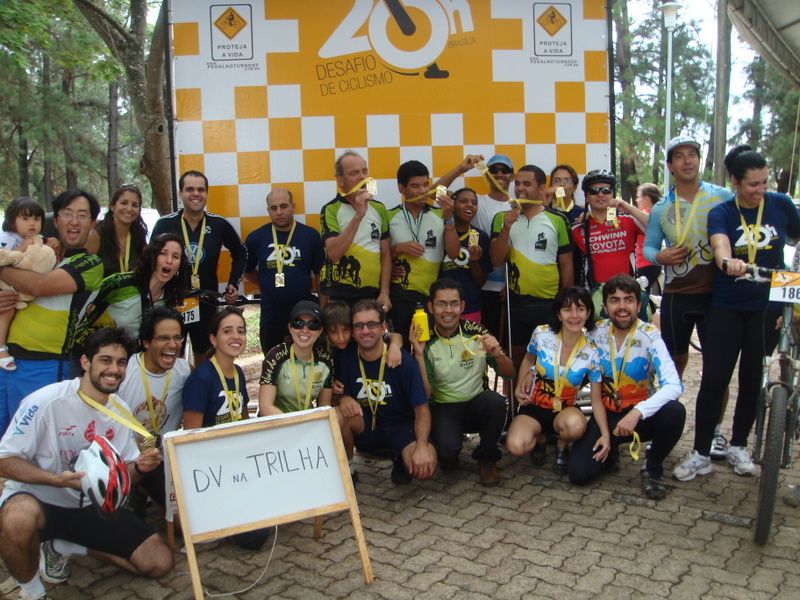 Imagem mostra organizadores do evento e todos os participantes do projeto DV na Trilha, com medalhas no pescoço em frente ao banner do evento.